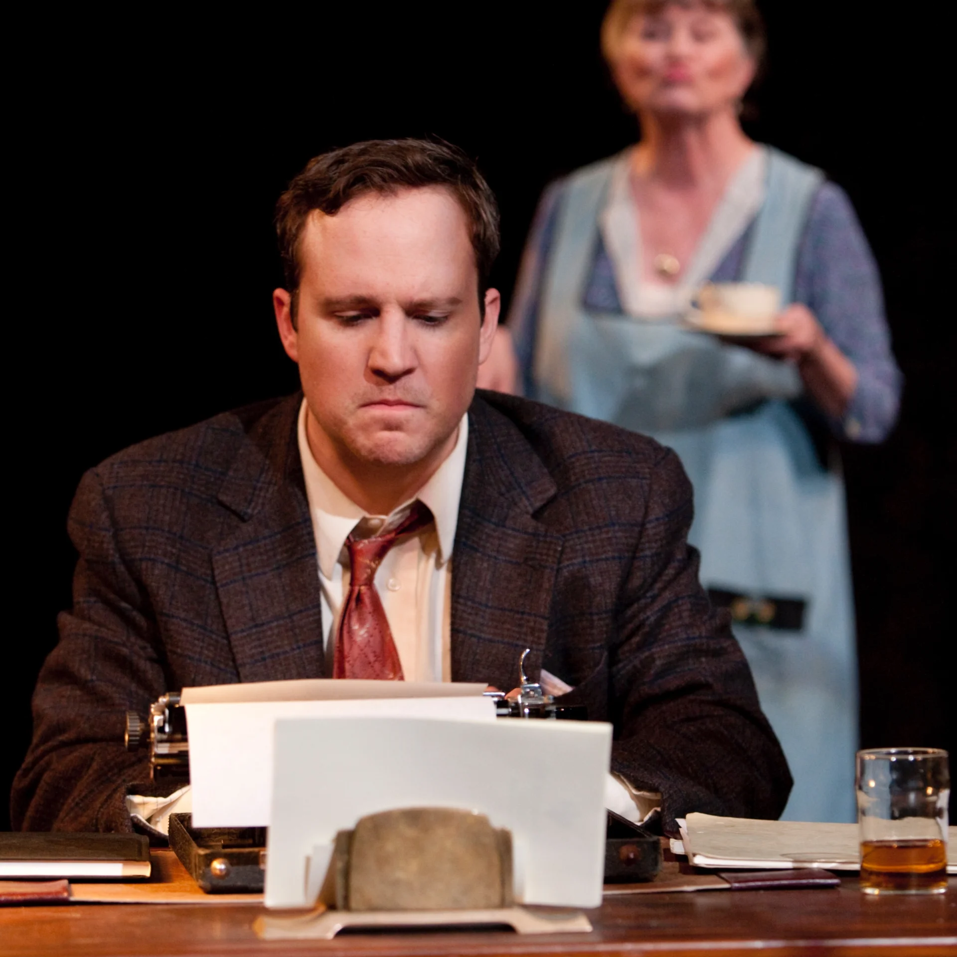 A man works at typewriter while a woman lingers in the background