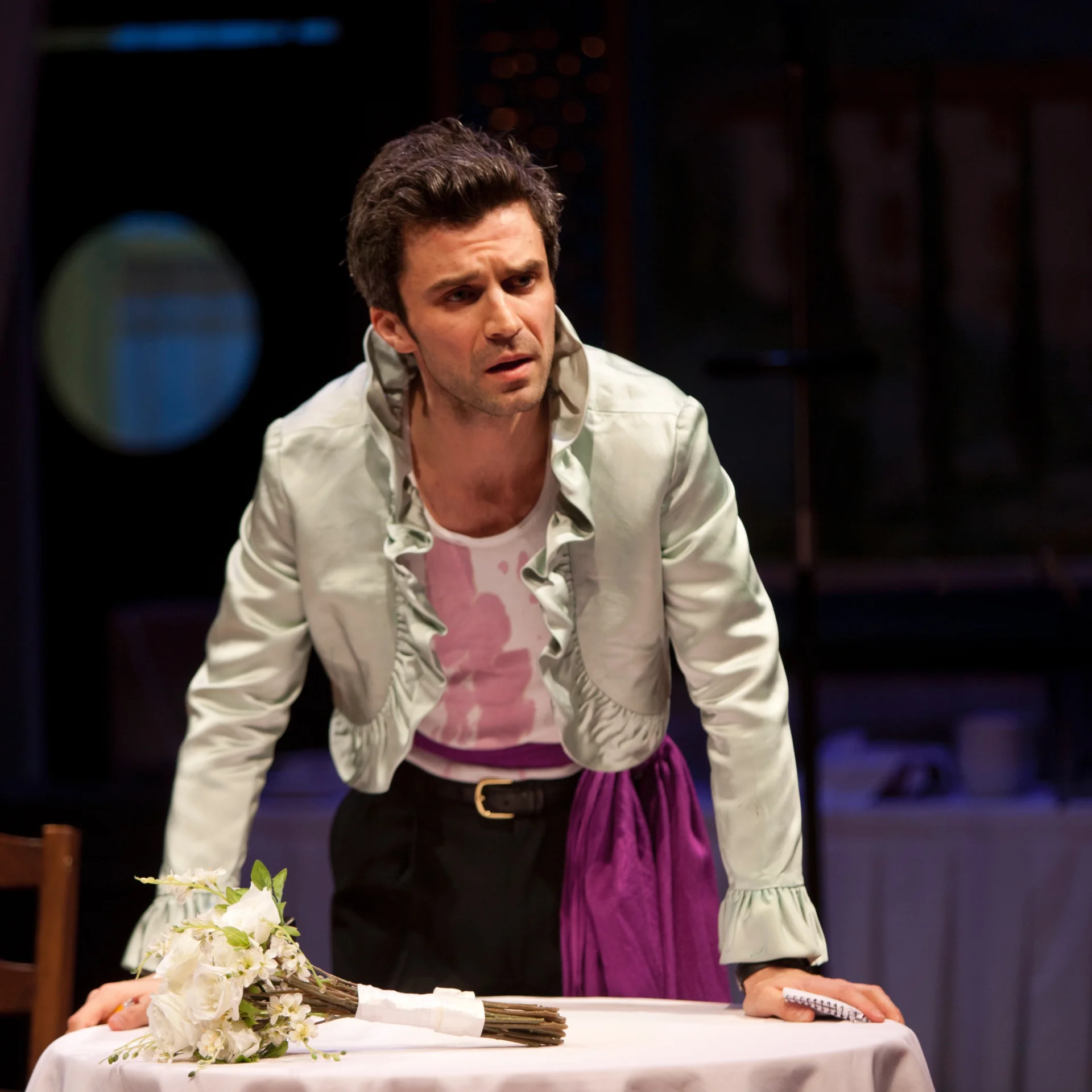 A man in a frilly shirt leans on a table with a bouquet of flowers