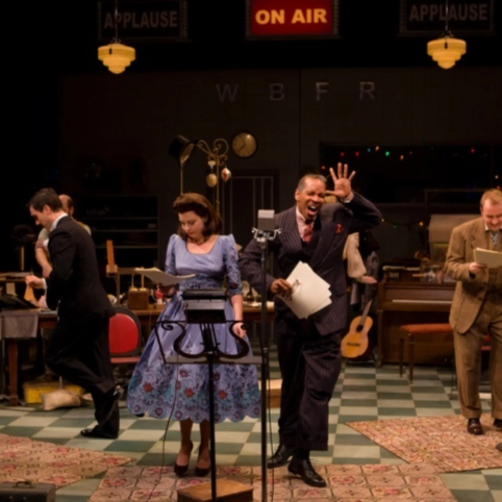 A woman in a blue dress and man in a suit stand in front of a microphone holding scripts in a studio