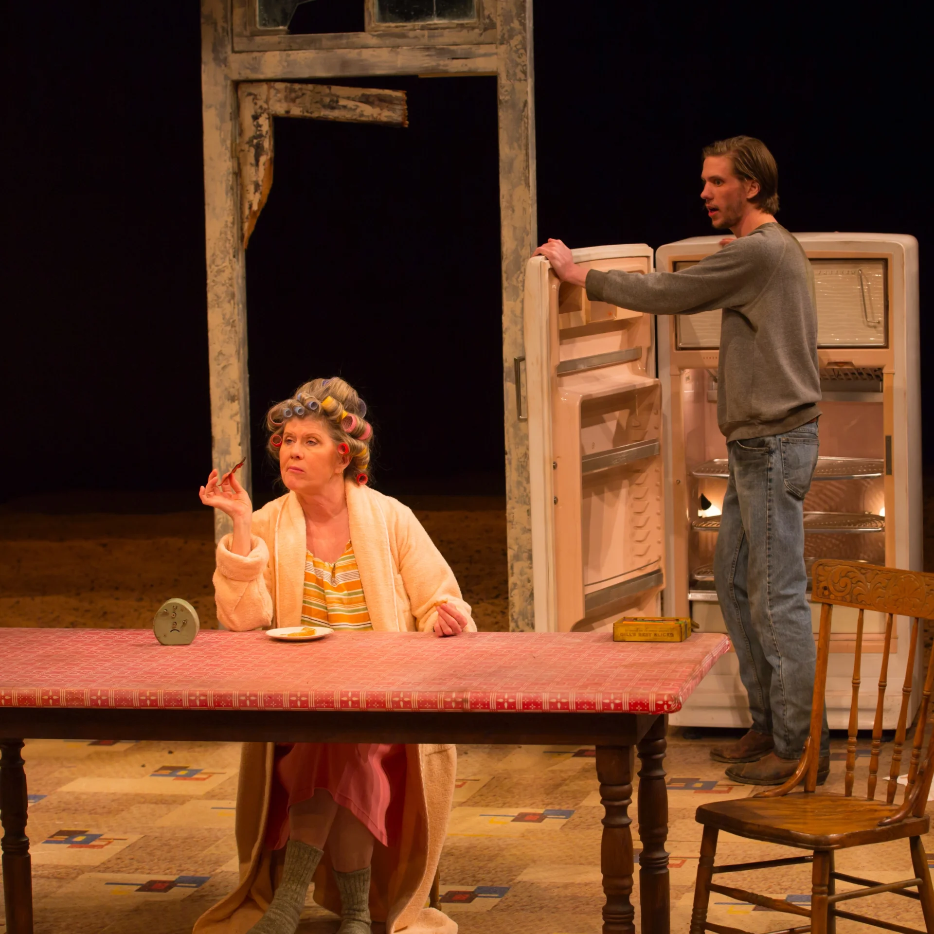 A woman with rollers sits at a table eating while a man stands opening a fridge behind her