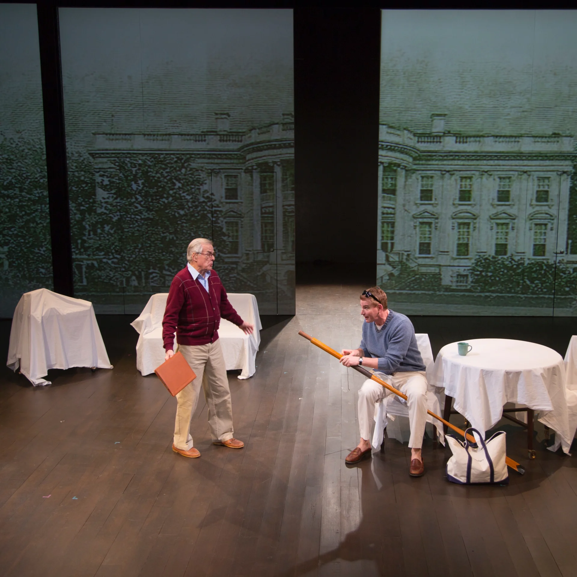 A man holding an oar sitting at a round table talks with another man who is standing