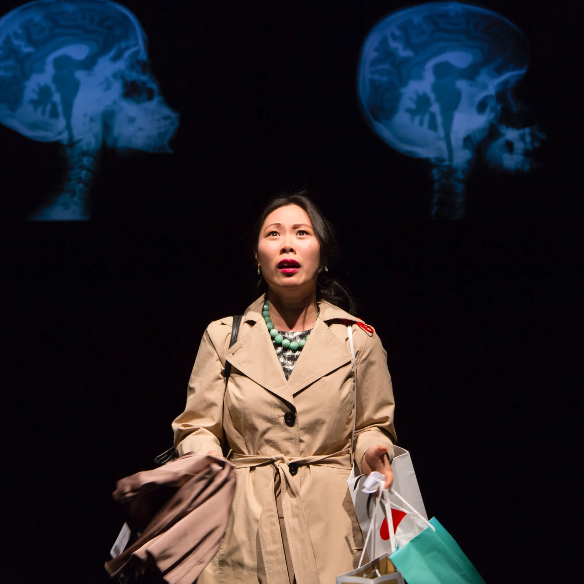 A woman in a trench coat looks out as two CT scans of skulls are projected behind her