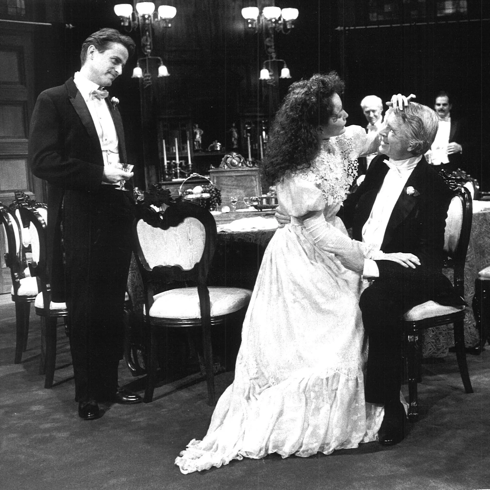 A woman is sitting on a mans lap wearing a long dress while a man in a suit is watching from behind