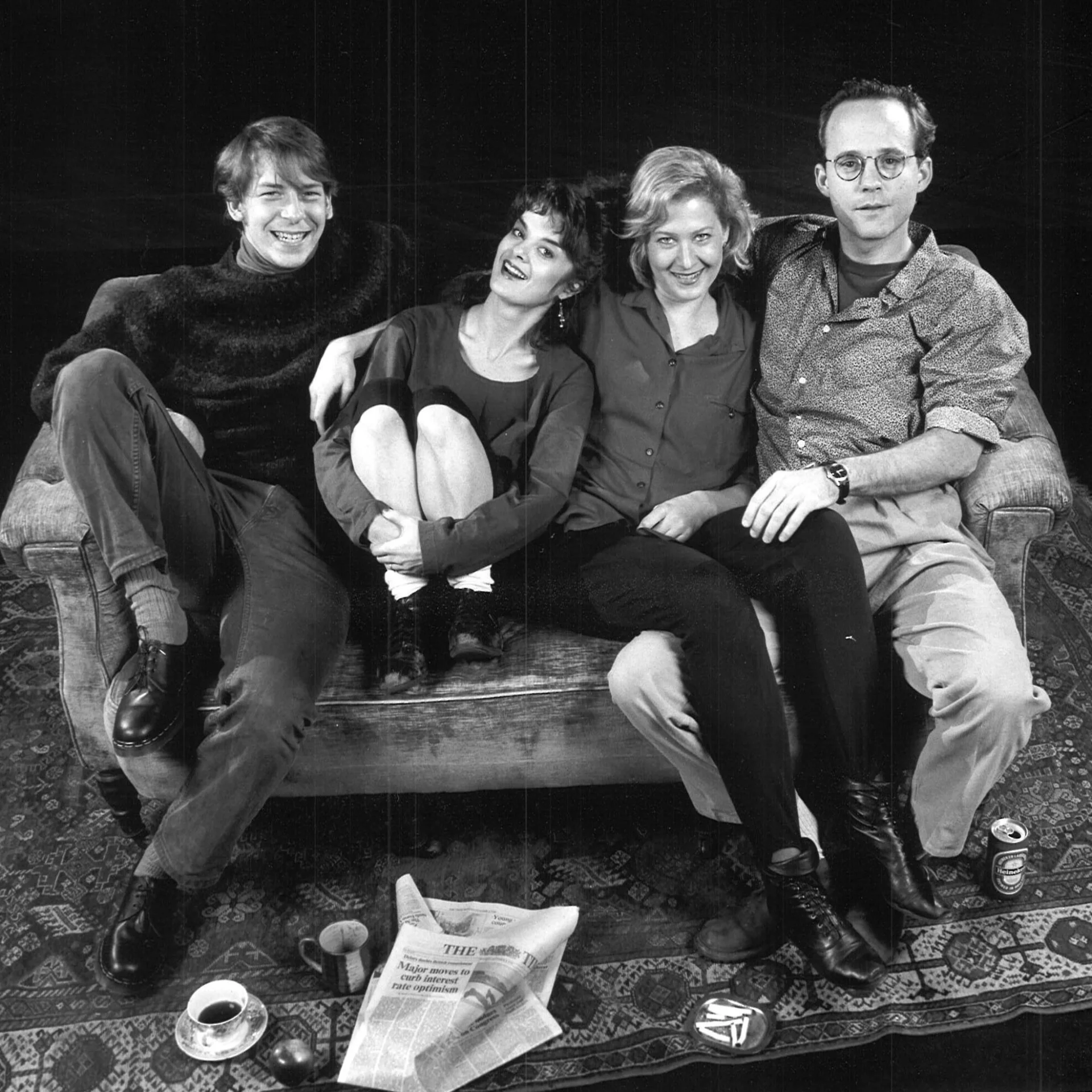 A group of people are sitting together on a couch and there is a newspaper and cup of coffee on the ground in front of them.