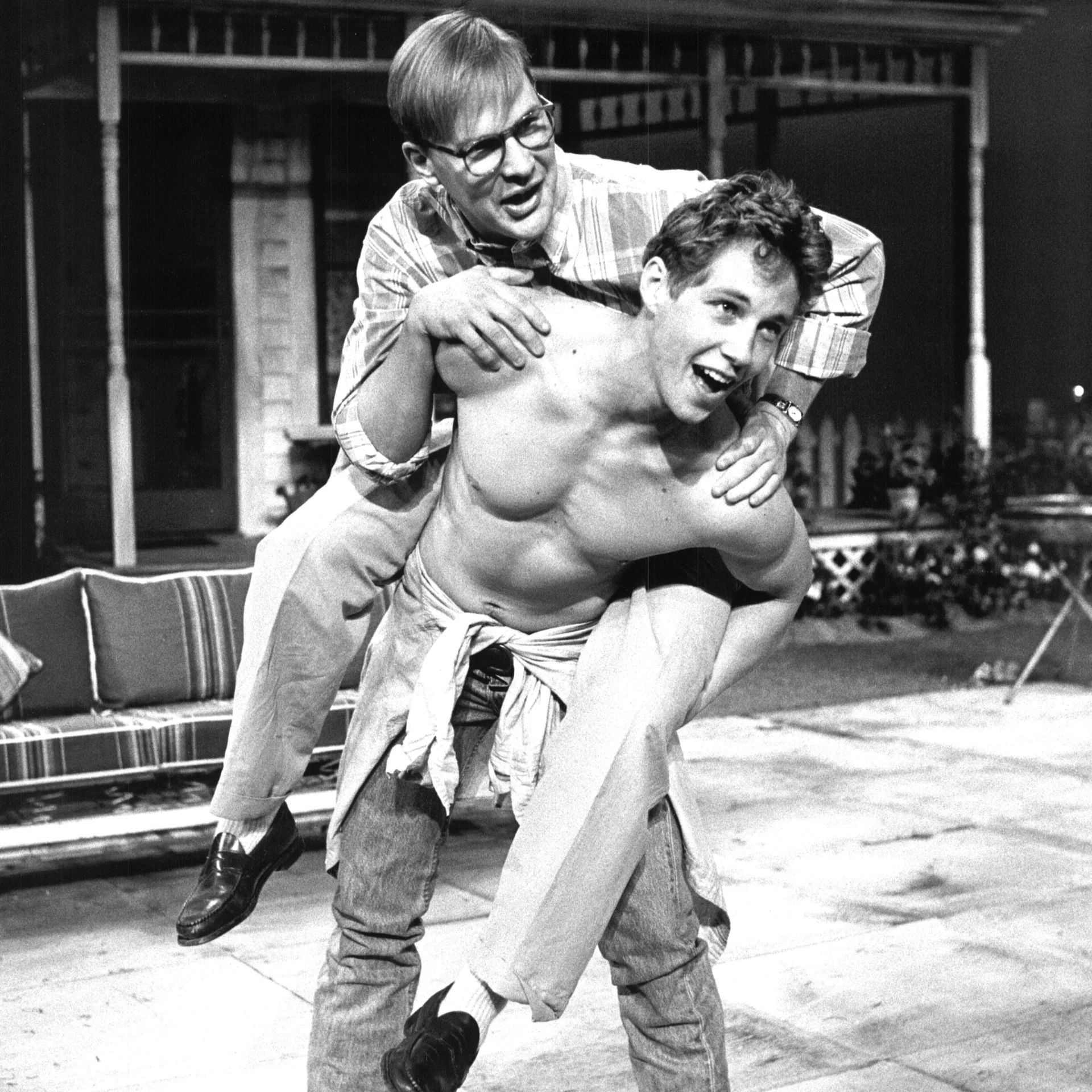 A Man wearing glasses is jumping on top pf a man who's shirtless and they are wrestling.