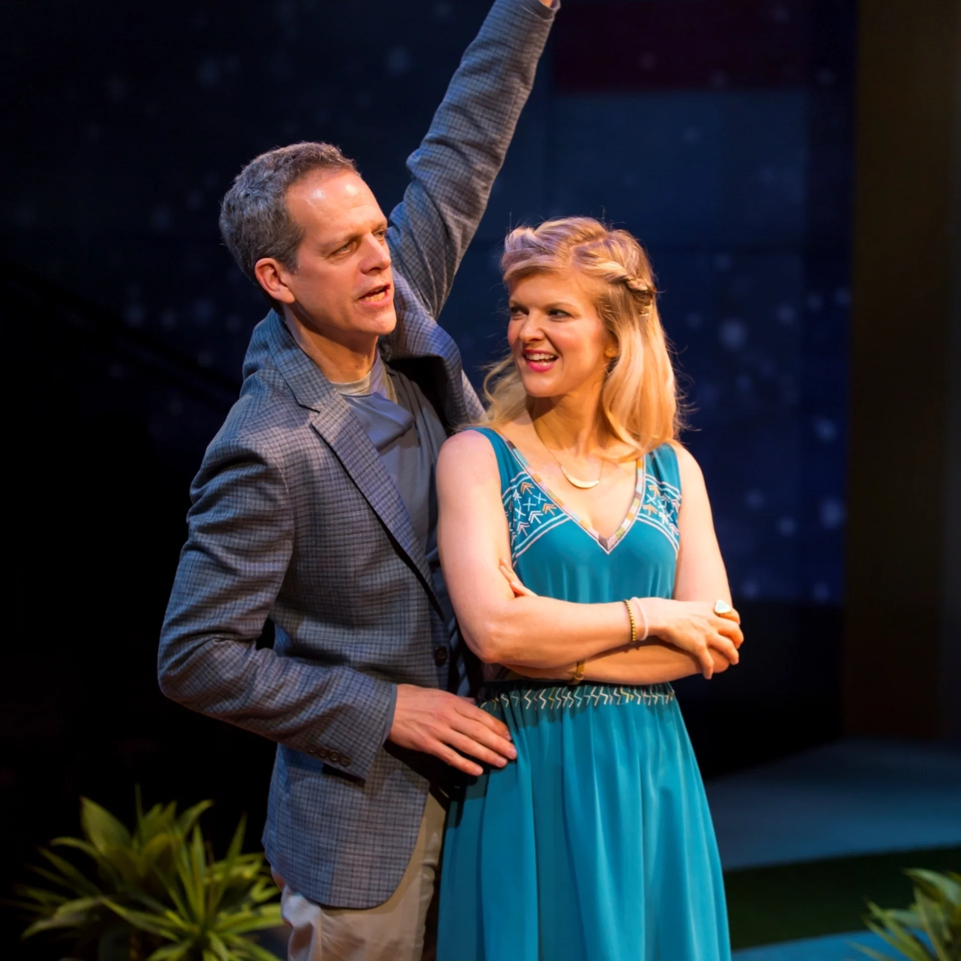 A man and woman stand on stage. The man is raising his left arm, while the woman stands beside him with her arms crossed and smiling. They are in a theatrical setting with plants in the background.